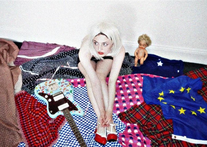 Girl in centre of image with blonde hair sitting on top of different blankets with a bass guitar to her left and a toy baby doll to her right.x